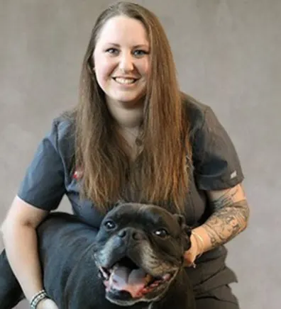 Heather Glick smiling in front of a grey backdrop hugging a black dog